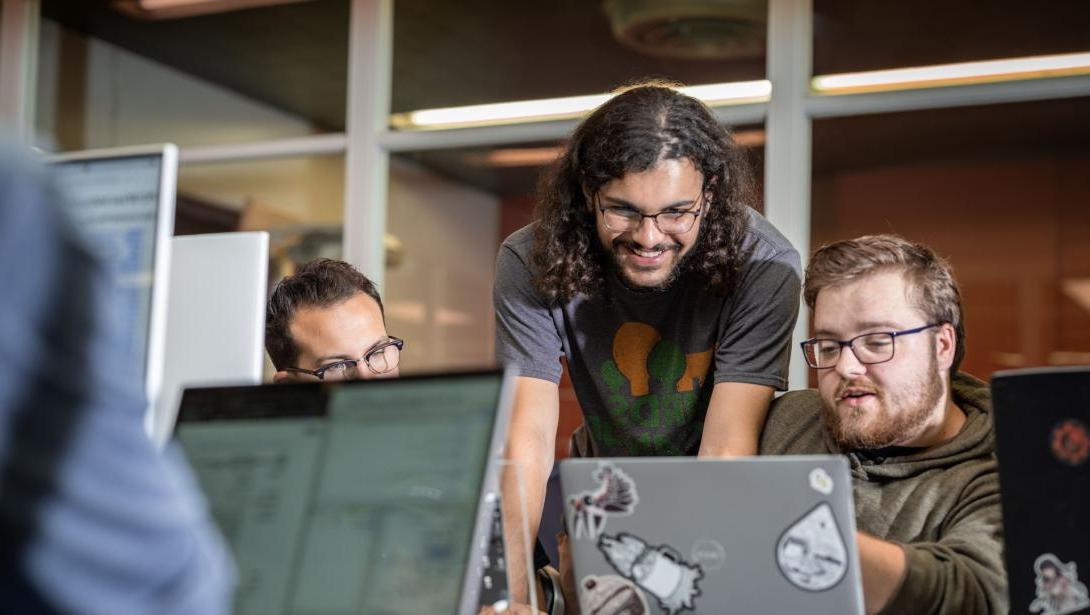 Students gathered around a table looking at and pointing to a laptop